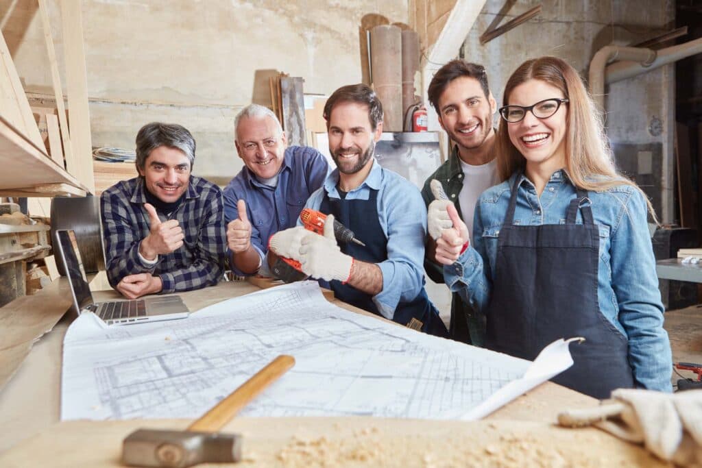 Fünf Handwerker in einer Werkstatt zeigen lächelnd den Daumen nach oben, vor ihnen ein Bauplan
