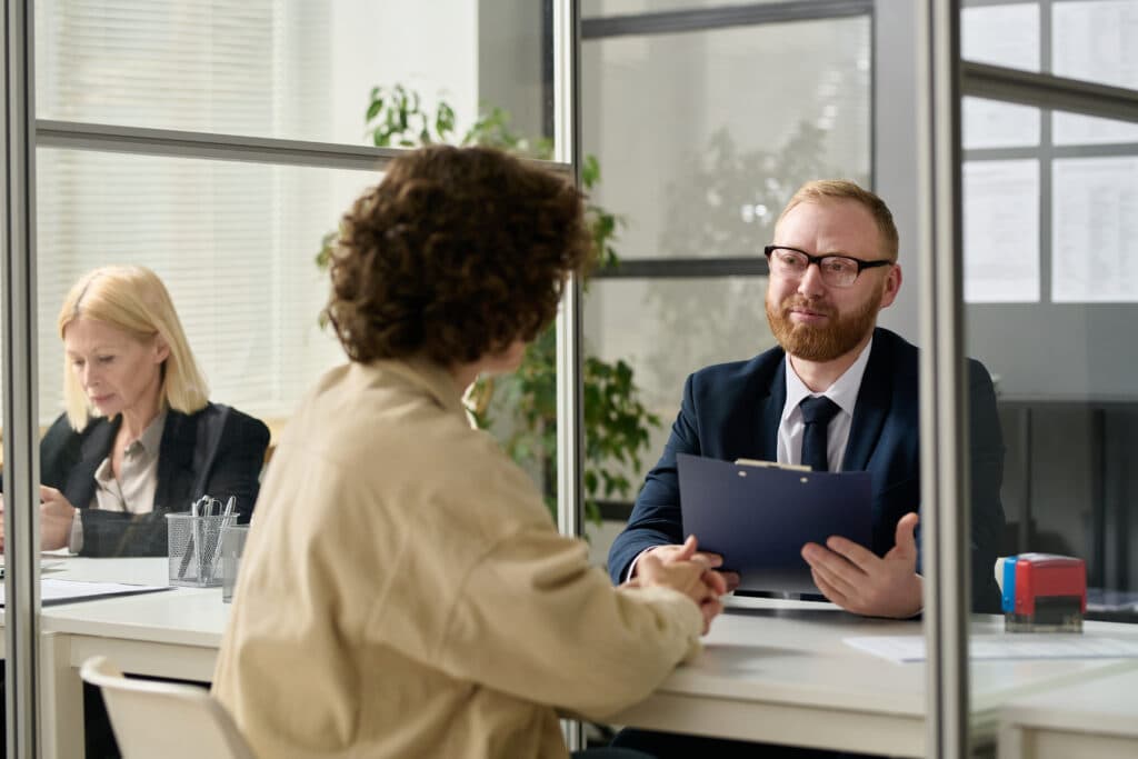 Bewerbungsgespräch im Büro: Ein Mann mit Klemmbrett spricht mit einer Bewerberin, im Hintergrund eine Kollegin