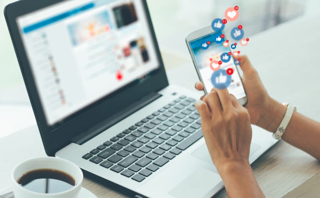Hände halten ein Smartphone mit aufsteigenden Like-Symbolen, daneben ein Laptop mit sozialem Netzwerk