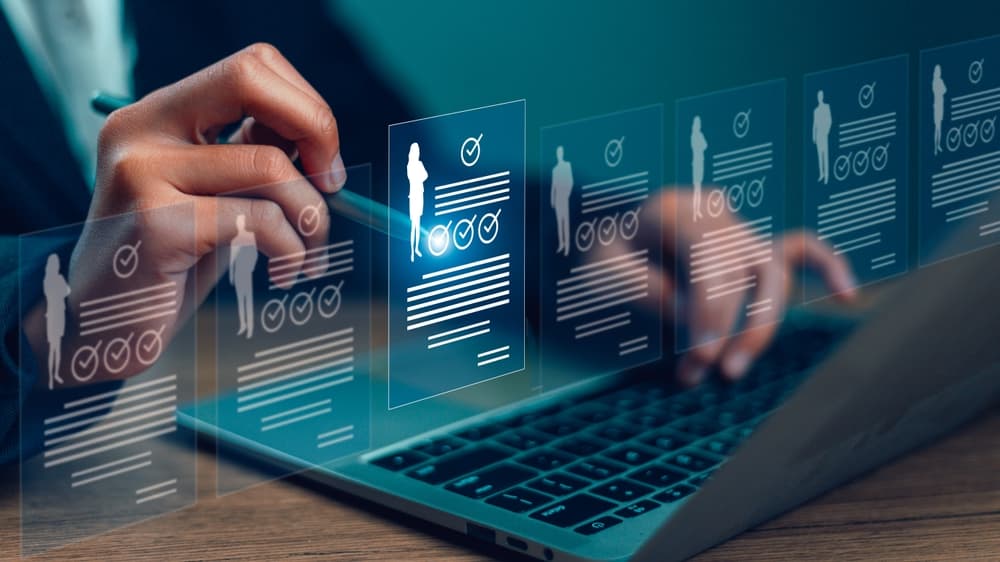 Hände an einem Laptop, darüber digital eingeblendete Bewerberprofile mit Häkchen