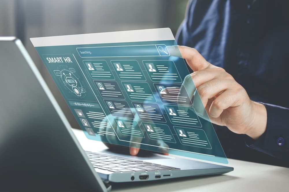 Hand an einem Laptop mit eingeblendeter digitaler Oberfläche „Smart HR“ voller Kandidatenprofile